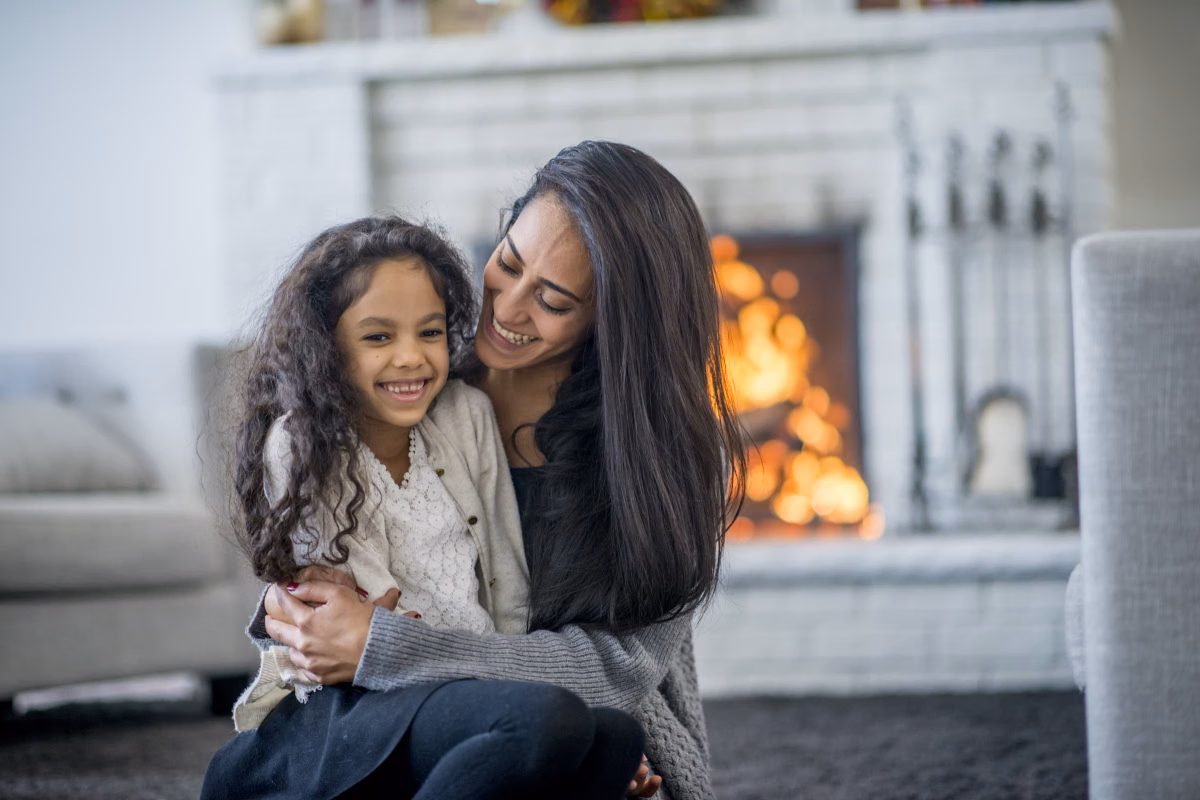 mother hugging her young daughter beside a warm open fireplace